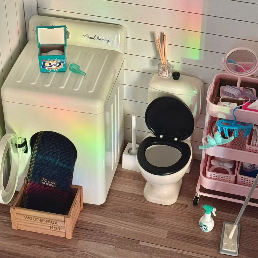 Open-door off-white miniature washing machine with doll clothes inside, beside a miniature toilet and pink trolley in a bathroom scene
