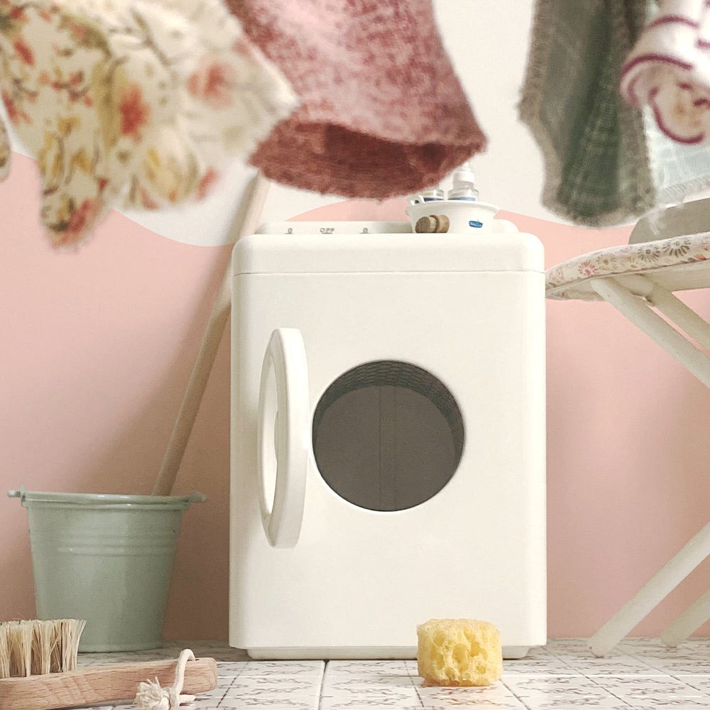 Close-up of an off-white miniature washing machine with front door open, hanging doll clothes and cleaning props in a pink laundry scene