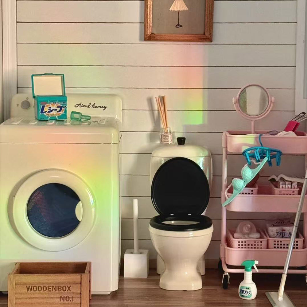 Off-white miniature washing machine with closed front door beside a miniature toilet and pink storage trolley in a dollhouse bathroom scene